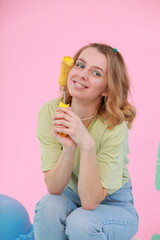 A young woman in a green T-shirt and jeans sits on a chair in a bright studio, playfully holding a large macaron-shaped pillow. Decor: pink background, big cactus, dessert-style props.