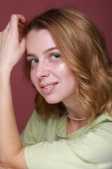 Fototapeta premium A portrait of a young blonde woman with wavy hair against a dark red background. She tilts her head, resting her hand. Her makeup features glitter on her lips and cheek. She wears a light green T-shir