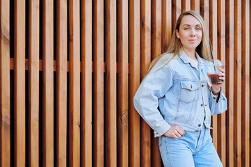 Young woman in denim jacket and jeans leaning casually against wooden slatted wall while holding a...