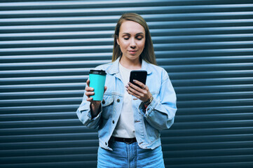 Stylish young woman in denim jacket holding reusable coffee cup and smartphone while standing...