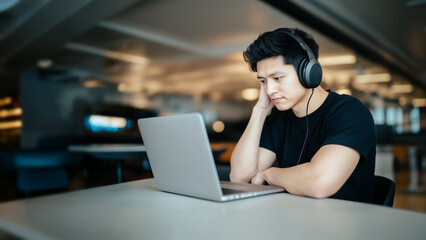 man wearing headphones and black T shirt is focused on his laptop in modern workspace. setting is well lit, with blurred background suggesting busy environment