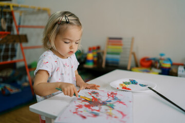 Little girl draws in an album with a brush with paints sitting at the table