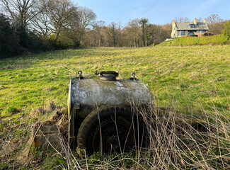 A rusty, cylindrical tank sits in a grassy field, partially hidden by tall grass. In the...