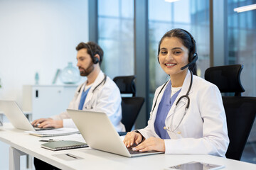 Smiling female doctor wearing a headset and stethoscope works on her laptop in a modern office.