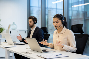 Two professionals are engaged in a video conference at their office workstations with headsets.