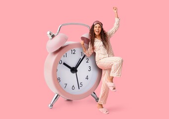Happy African-American woman in pajamas and big alarm clock on pink background