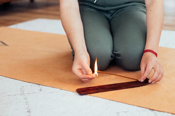 A close-up of a woman sitting on a yoga mat, lighting an incense stick for meditation and relaxation. Cozy home atmosphere with warm tones and a peaceful vibe.