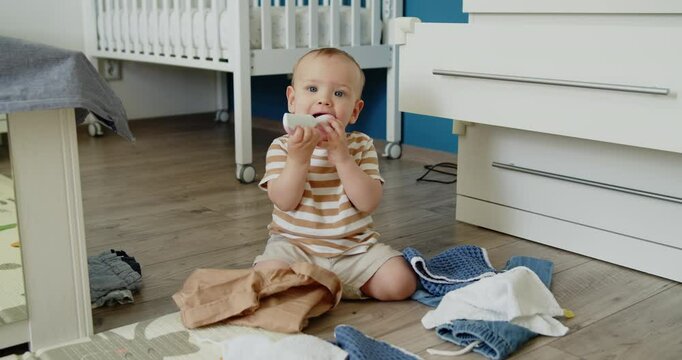 Baby teething. A one year old child gnaws a comb sitting on a bedroom floor surrounded by clothes taken out of a dresser. Slow motion middle shot.