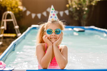Blonde 8-year-old girl swimming in a home pool during a summer party, wearing blue swimming goggles and a paper birthday hat
