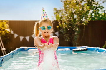 Red haired 7-8 year old girl celebrating her birthday in a summer pool, jumping in the water and...