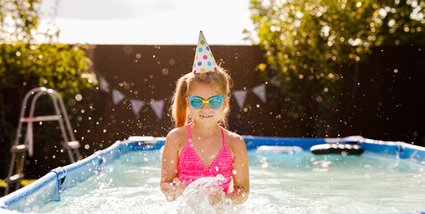 Red haired 7-8 year old girl celebrating her birthday in a summer pool, jumping in the water and...