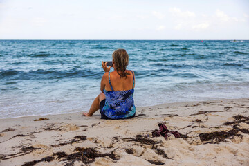 Woman in blue swimsuit sitting on sandy Miami Beach, taking photo of Atlantic Ocean waves under cloudy sky. USA.