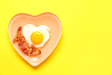 Plate with tasty bacon and heart made of fried egg on yellow background