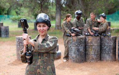 Fototapeta premium Smiling asian woman paintball player in camouflage posing with gun outdoors