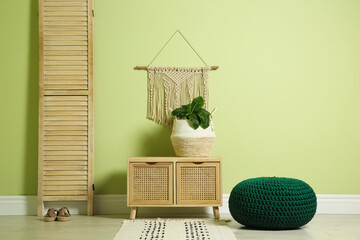 Stylish room interior with comfortable knitted pouf, wooden furniture and plant near light green...