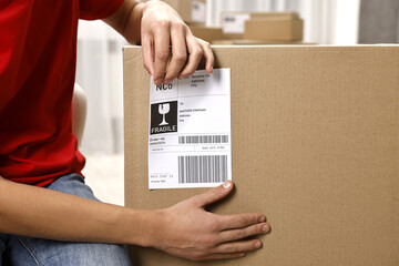 Man sticking shipping label with barcode on parcel indoors, closeup