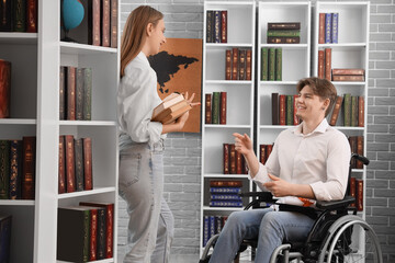 Male graduate in wheelchair talking to his friend with books at library