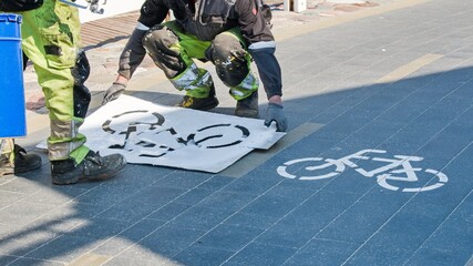 Workers in paint-stained overalls renew the marking of the bicycle path in the spring with the help of a stencil and a spray bottle