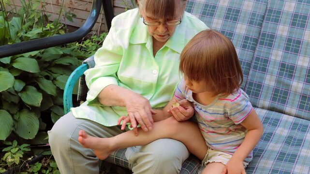 A caring grandmother sits on an outdoor cushioned sofa, gently applying ointment to her young granddaughters leg to soothe a mosquito bite