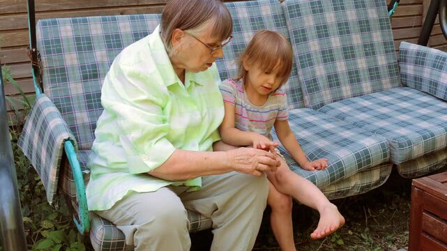 A caring grandmother sits on an outdoor cushioned sofa, gently applying ointment to her young granddaughters leg to soothe a mosquito bite