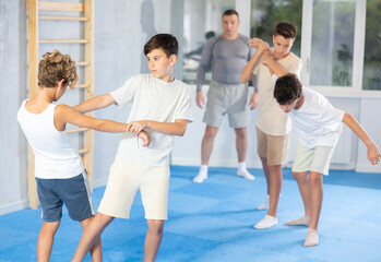 Focused teenage boys practicing close combat techniques in sparring in training room during self-defence workout under supervision of coach