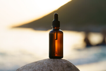 Brown mockup of a glass jar of serum with a dropper on the stone against the background of the sunset sea and rays of the sun. Concept of beauty products, skin care