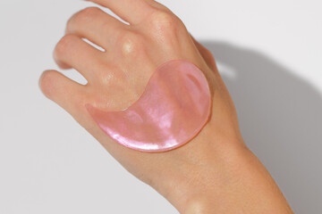 Close-up of a pink hydrogel eye patch on female hand skin against a white background. Hydrates,...