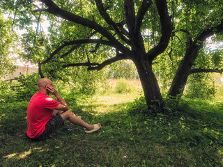 A man in a red shirt stands in shade in front of a tree. The image has a peaceful and serene mood, as the man is standing in a lush green garden on a hot summer day. Country life and holiday theme.