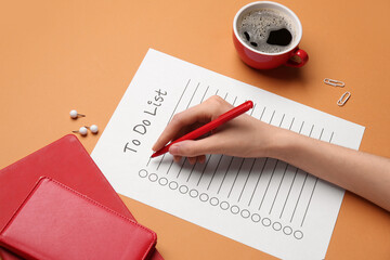 Female hand with to do list, cup of coffee and stationery on orange background. Closeup
