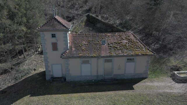 An old beige building with a moss-covered tiled roof stands in a rural setting. It has a small tower, closed windows, and a wooden door. Shadows from the sun highlight its aged appearance and worn str