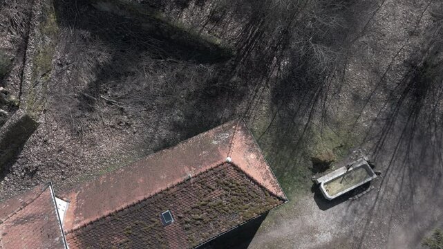 An old beige building with a moss-covered tiled roof stands in a rural setting. It has a small tower, closed windows, and a wooden door. Shadows from the sun highlight its aged appearance and worn str