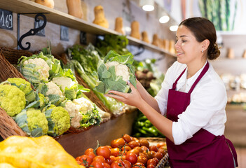 Positive young shop assistant putting fresh cabbages on counter in large greengrocery
