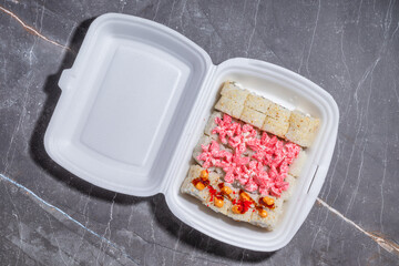 A top-down view of a sushi takeout box with different sushi rolls arranged neatly. The rolls include creamy seafood topping, spicy mayo, and sesame seed-covered sushi.
