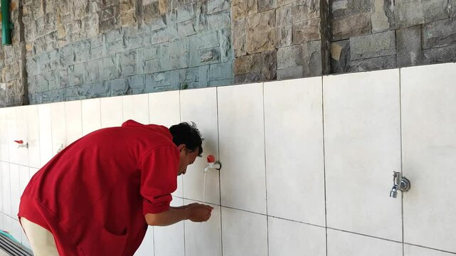 An Asian Muslim man wearing a red jacket is performing ablution (cleansing oneself with water) before praying.