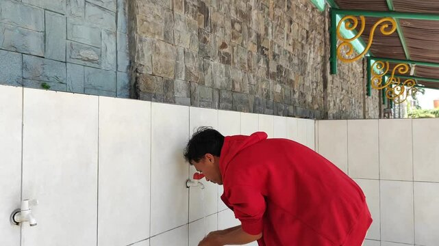 An Asian Muslim man wearing a red jacket is performing ablution (cleansing oneself with water) before praying.