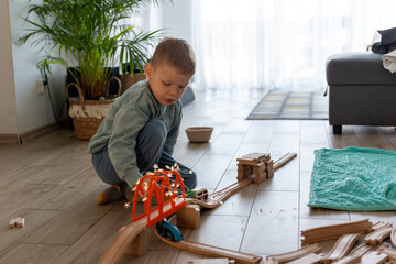 Little boy in cozy home environment playing with wooden train set on the floor, carefully arranging...