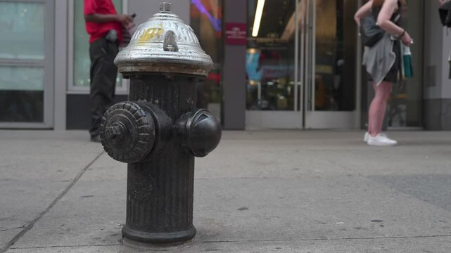 Crowds of pedestrians navigate a bustling Manhattan sidewalk. A classic black fire hydrant stands prominently as people pass by, showcasing city life.