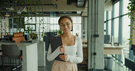 smiling young professional woman with glasses holding a clipboard in a modern office space with...