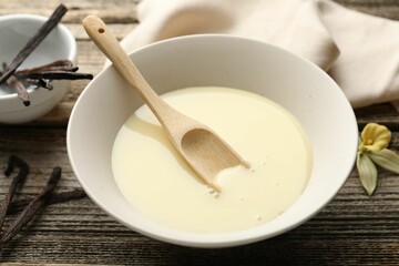 Tasty condensed milk, spoon, vanilla pods and flower on wooden table, closeup