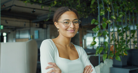 A confident, smiling young professional woman with glasses and arms crossed, standing in a modern,...