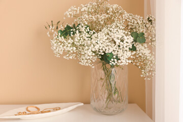 Bouquet of beautiful gypsophila flowers and eucalyptus branches in vase on chest of drawers near beige wall