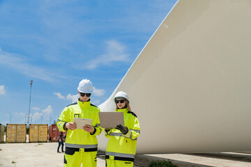 Two engineers in high visibility gear use a laptop to work in front of a large wind turbine blade at a construction site. the integration of technology in renewable energy and wind power projects.