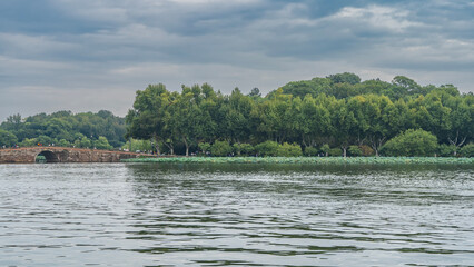 Beautiful calm urban lake. Lotuses near the shore. People are walking in the park among the green trees. Stone arched bridge over the water. Blue sky, clouds. China. Hangzhou. West Lake Xi Hu
