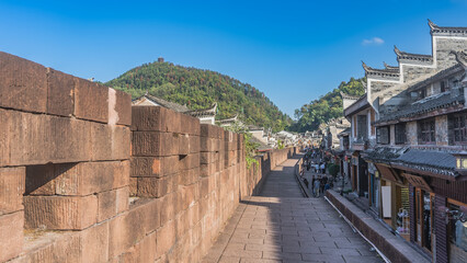 Fototapeta premium The ancient brick fortress wall of Fenghuang Old Town. Traditional Chinese buildings along a paved street. People are walking on the sidewalk. The mountain against the blue sky. China.