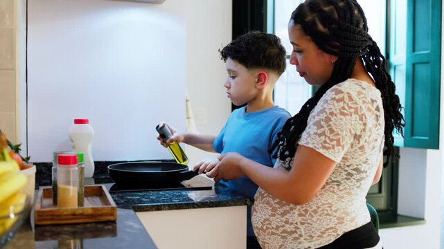 Pregnant latin mom and son preparing a meal together in a modern kitchen, pouring oil into a frying pan