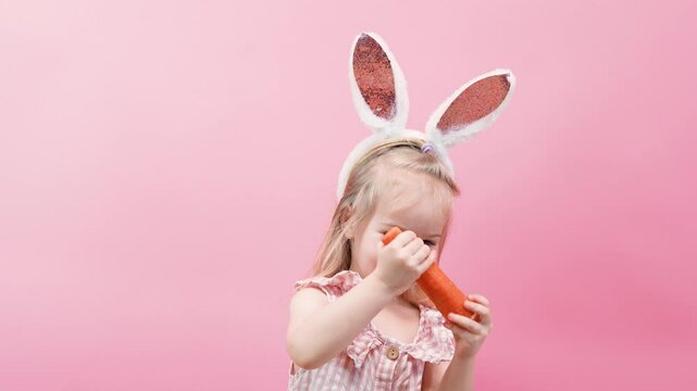 Cute Little child girl toddler nibbles a carrot like a bunny on an isolated pink background