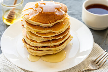 Pancakes poured with honey on a white plate with napkin and coffee on a gray background