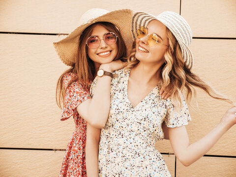 Two young beautiful smiling female in trendy summer sundress. Carefree women sitting at the stairs on the street background. Positive models having fun and hugging