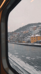 Train window view of a snowy riverside town with colorful houses on a hillside.