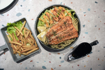 Grilled salmon served on a bed of pasta with fresh salad and lemon, paired with crispy fries and a side of savory black pepper sauce.